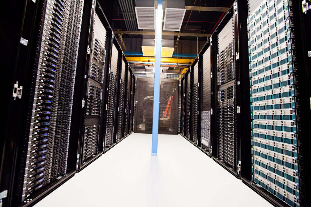 An aisle between tall server racks, with overhead cooling equipment and a central support column.